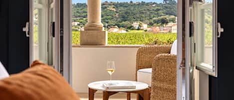 A room with a bed, a round table, a glass of wine, and wicker chairs.