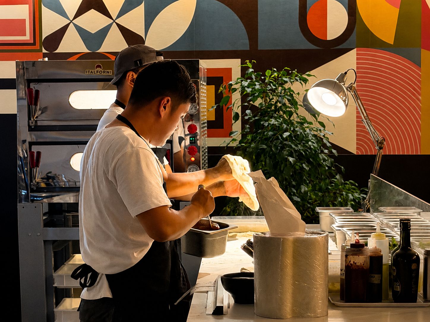 Une personne se trouve dans une cuisine, en train de préparer des aliments. On aperçoit en arrière-plan un four à pizza et divers ustensiles de cuisine.