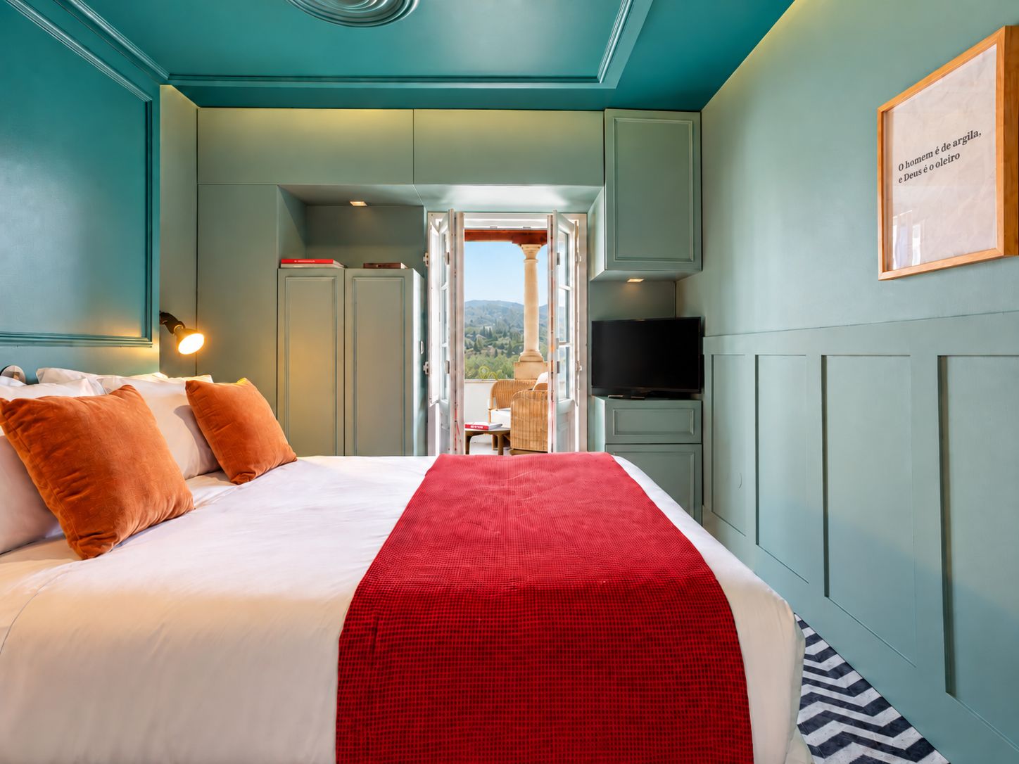 Habitación de hotel con cama, televisión, una manta roja y vista al exterior.