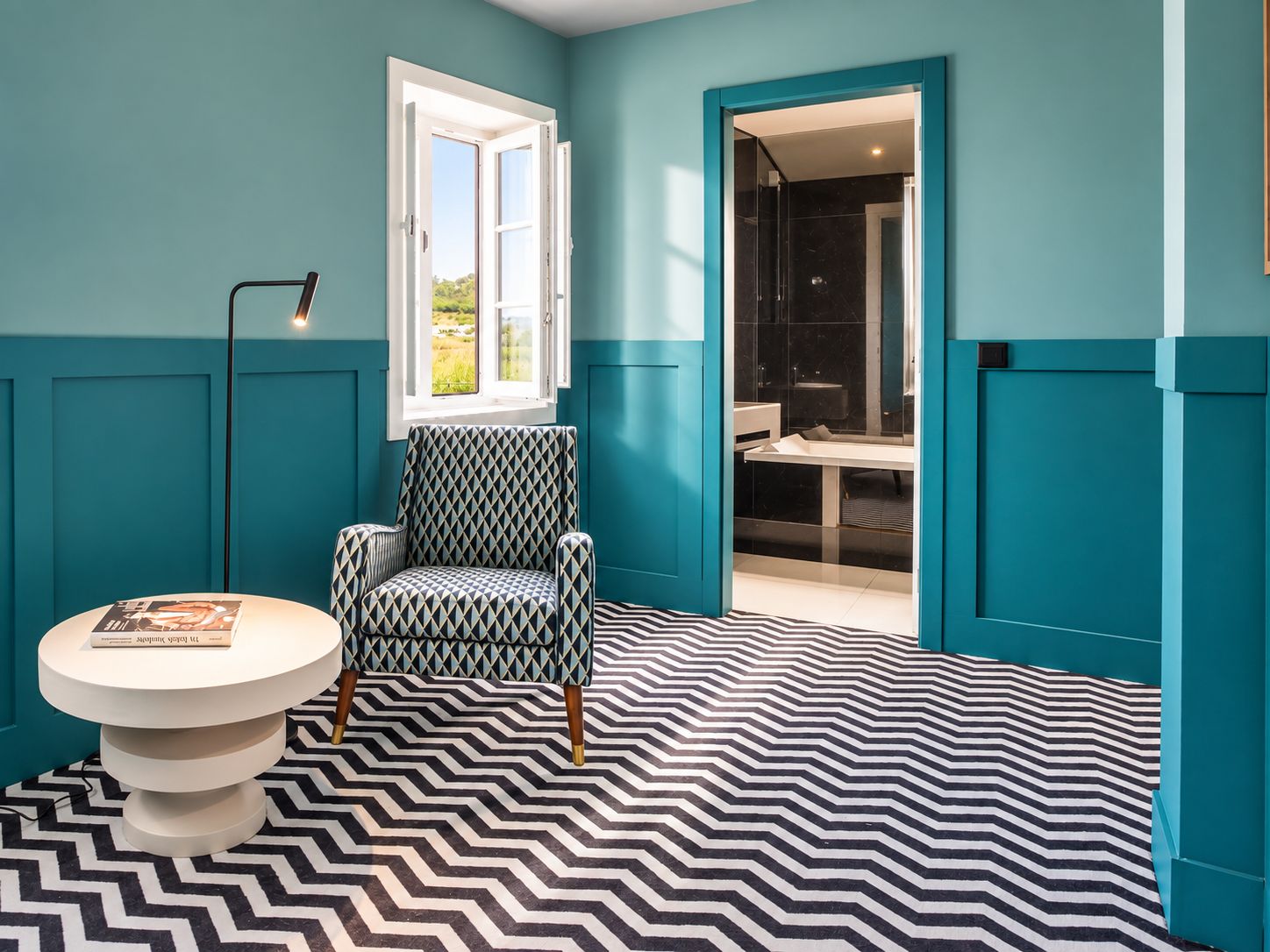Una habitación con paredes color turquesa, suelo estampado, una silla, una mesa con un libro y una ventana con vistas.