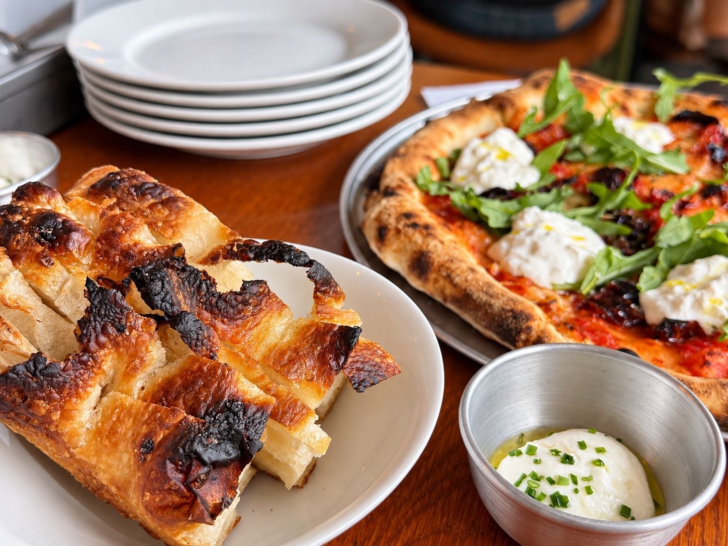 A table with a plate of grilled cheese, a pizza with toppings, and a small bowl of sauce.