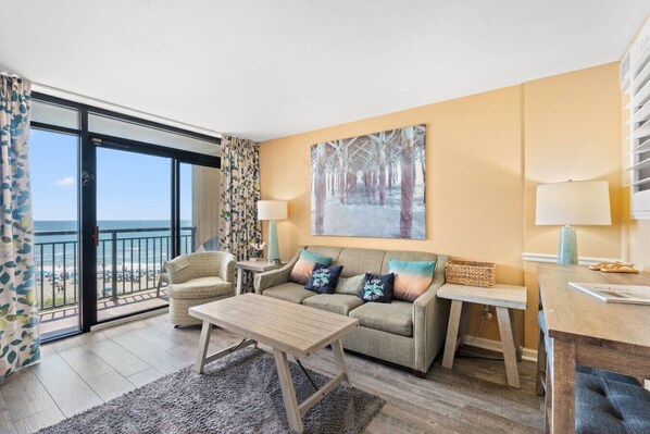 A hotel room with a balcony, a sofa, a coffee table, a side table, and a painting. - Oceanfront Condo Stay with Heated Pool and Hot Tub (North Myrtle Beach)
