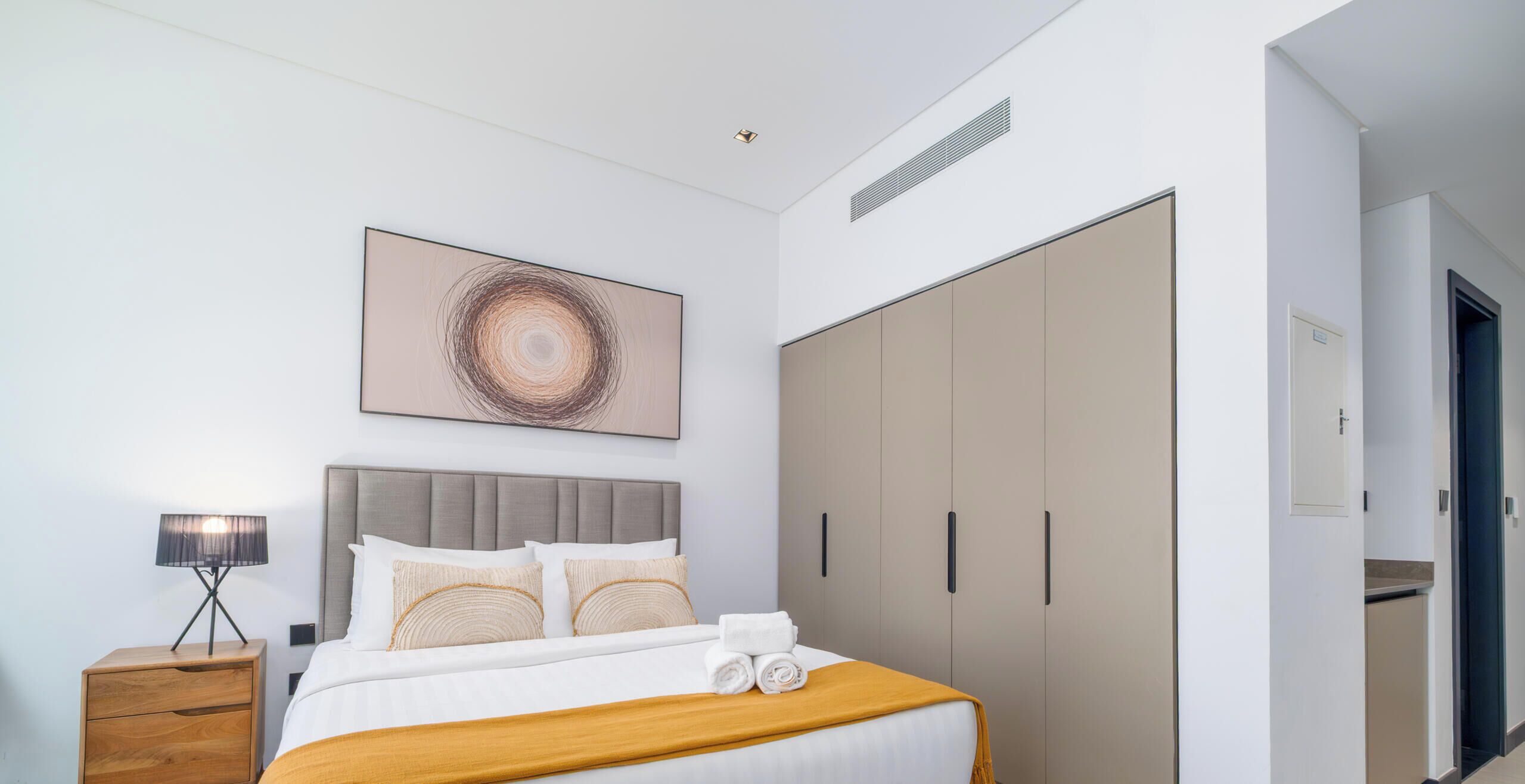 A modern hotel bedroom with a large bed, a bedside table, a wardrobe, and a wall-mounted artwork.