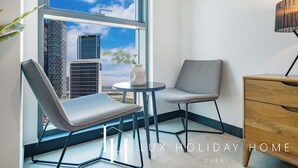 Een moderne kamer met uitzicht op de skyline van Dubai, met twee grijze stoelen, een klein rond tafeltje en een houten kastje.