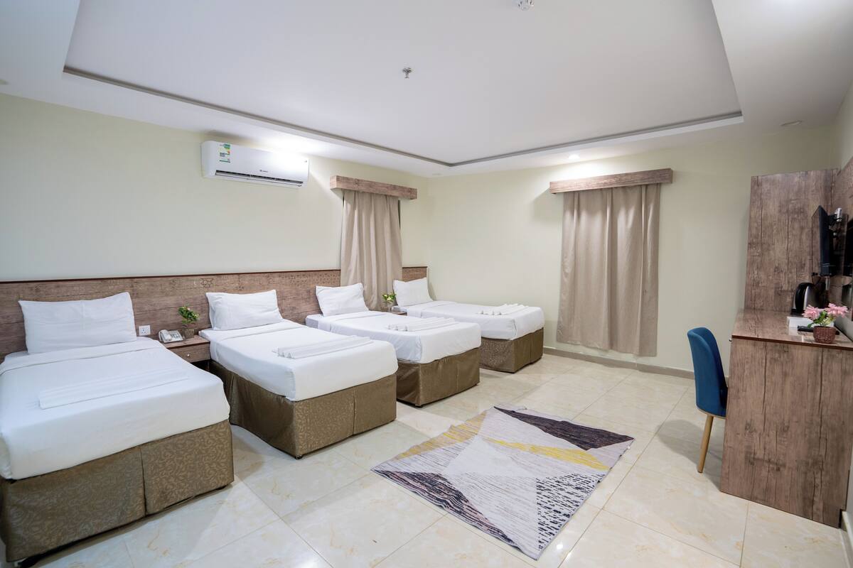 A hotel room with three single beds, a desk with a chair, a small table, and a large window with curtains.