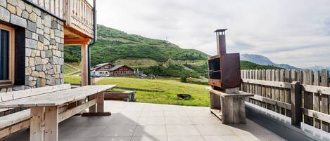 A wooden deck with a stone wall, a wooden bench, a grill, and a view of a house in a hilly area.