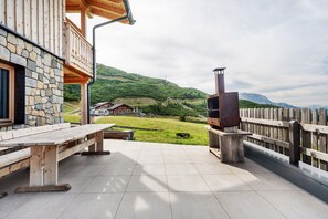 Una terraza de madera con muro de piedra, un banco de madera, una parrilla y vistas a una casa en una zona montañosa.