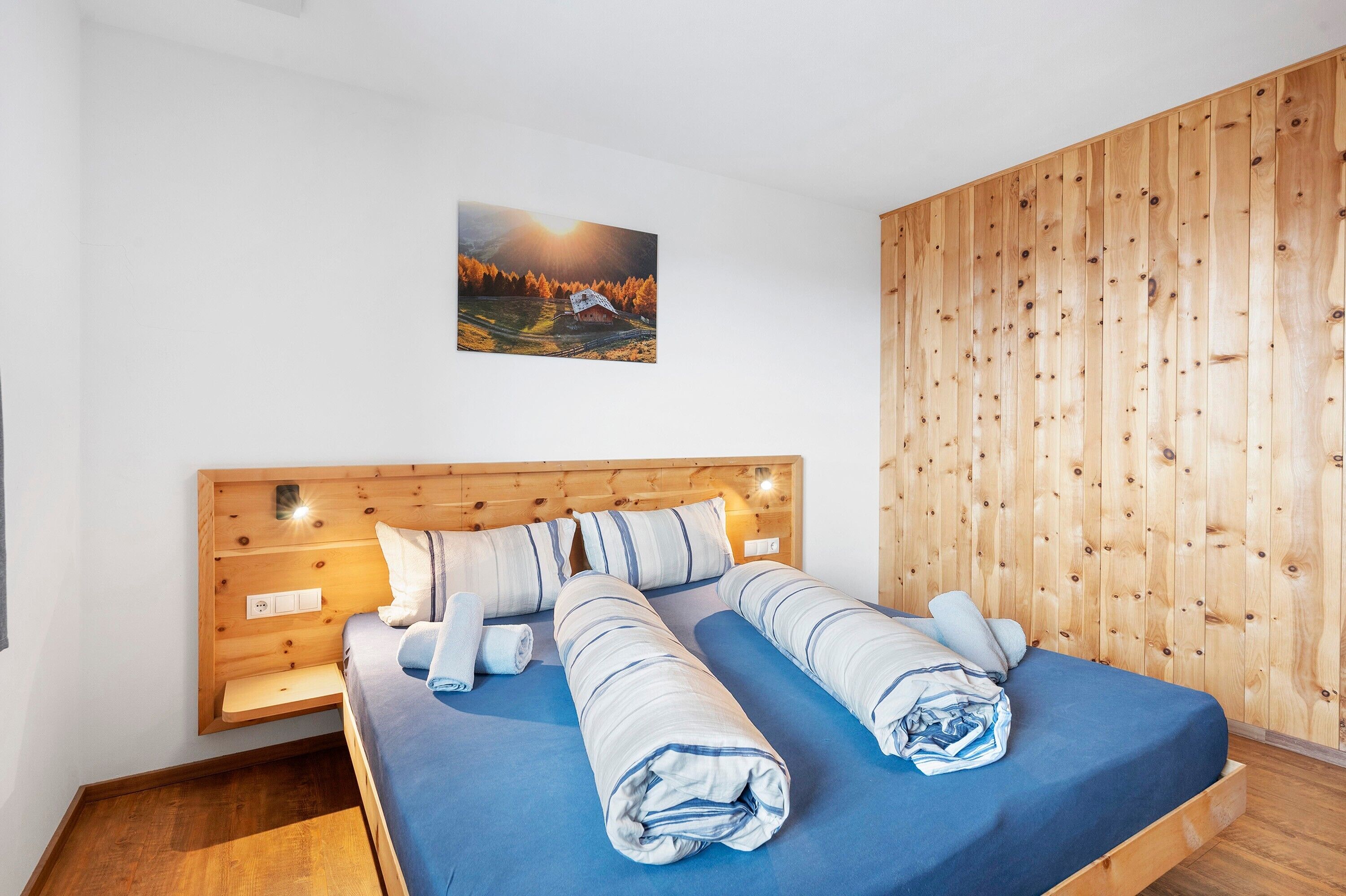 A bed with a wooden headboard, a painting above it, and a wooden panel wall.