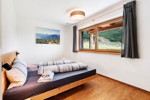 Una cama individual con cabecera de madera, vista a un paisaje montañoso por la ventana y una lámpara en el techo.