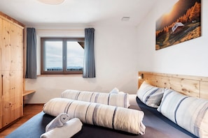 A bedroom with a wooden wardrobe, a large window with curtains, a bed with striped bedding, and a painting of a mountain landscape.