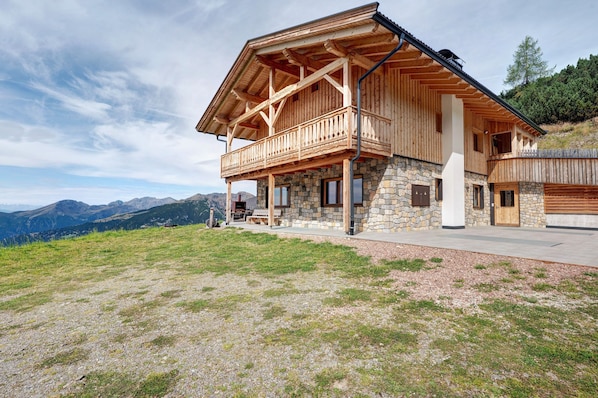 A wooden chalet with a balcony, stone foundation, and a view of mountains.