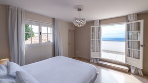 Quarto com cama, um lustre, janela com cortinas e varanda com vista para o mar.