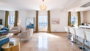 Sala de estar moderna com sofá, poltrona e mesa de centro. Há uma área de jantar com balcão e cadeiras. Uma grande janela oferece vista para o exterior.