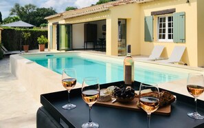 Escena junto a la piscina con una mesa dispuesta para una cata de vinos, incluyendo una botella, copas, queso y uvas.