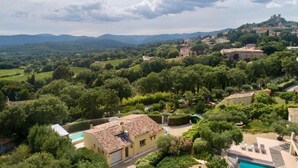 Una villa con tejado de baldosas, rodeada de exuberante vegetación y viñedos, con vistas a un paisaje montañoso y un castillo a lo lejos.