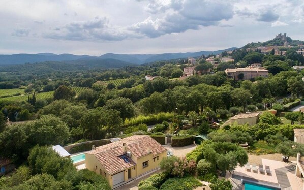 Una villa con tejado de baldosas, rodeada de exuberante vegetación y viñedos, con vistas a un paisaje montañoso y un castillo a lo lejos.