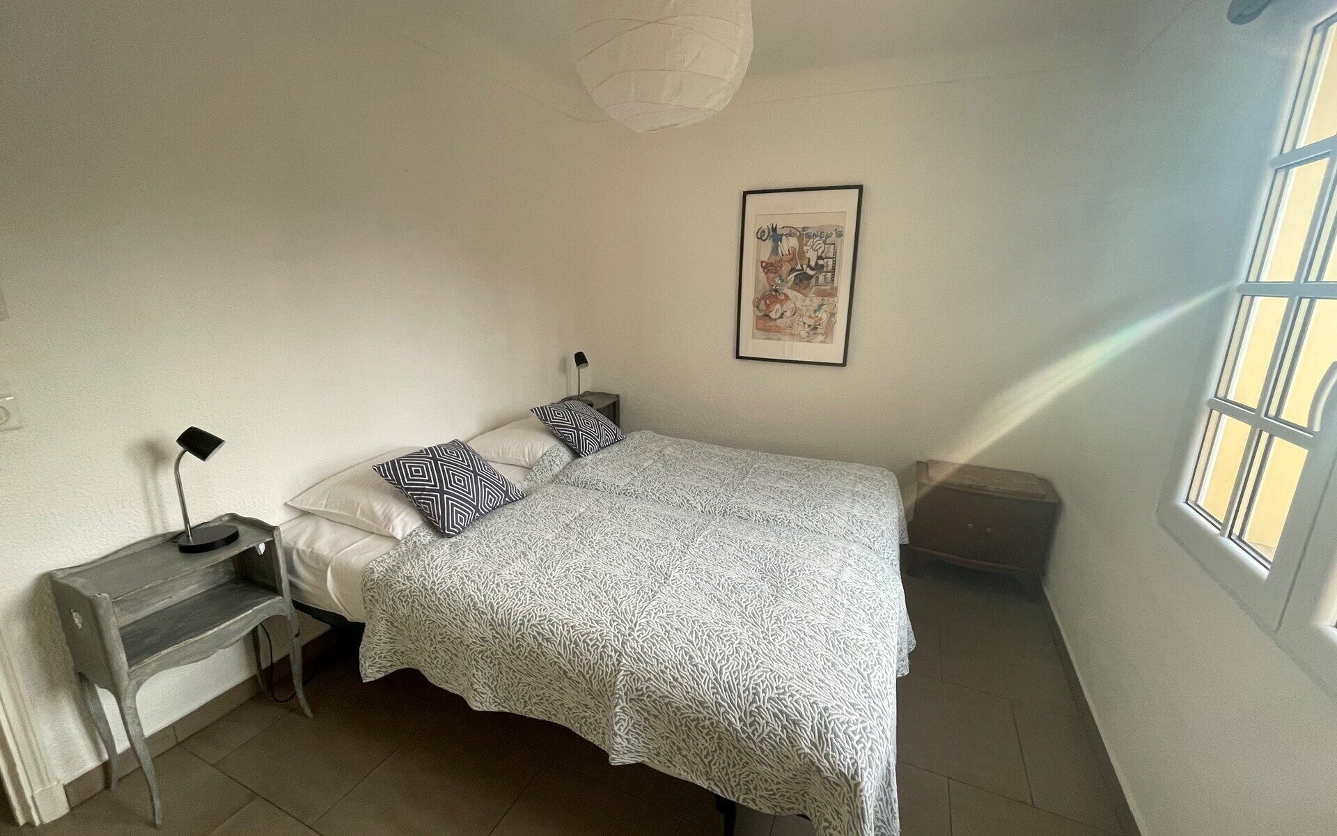 Un dormitorio con cama, escritorio, silla, lámpara, un cuadro en la pared y ventana.