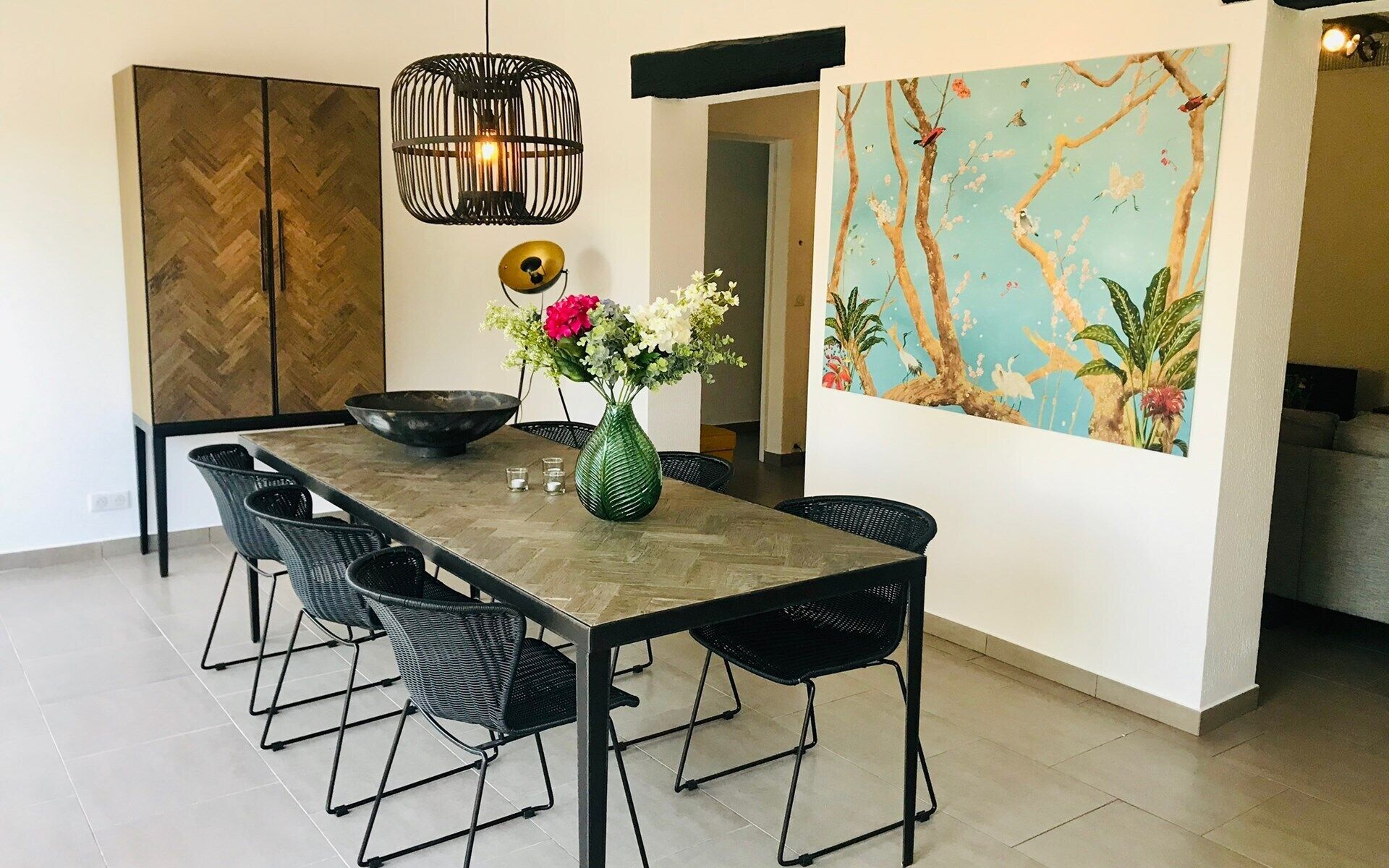 Een eetruimte met een lange tafel, zwarte stoelen, een groene vaas met bloemen, een hanglamp en een groot schilderij aan de muur.