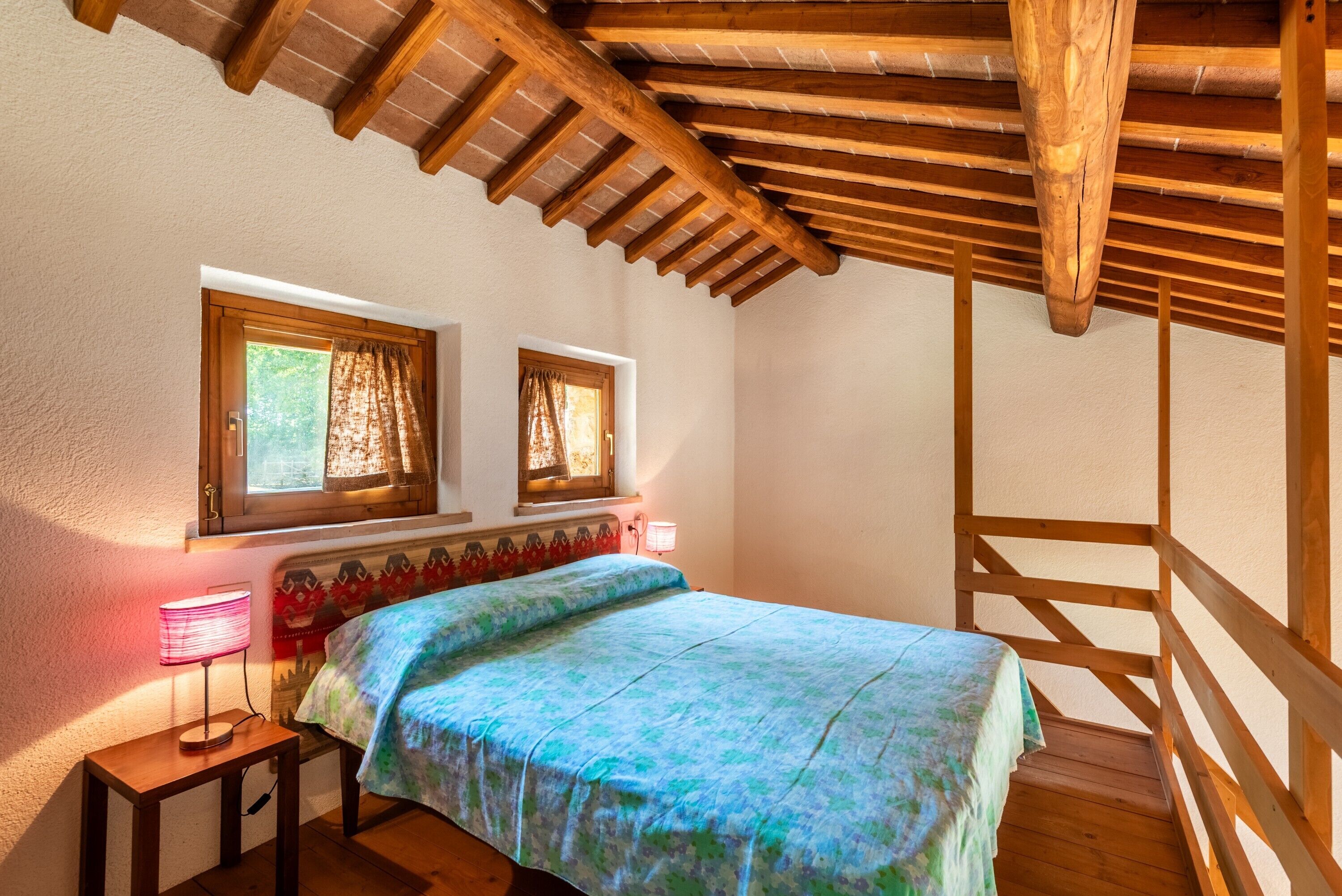 Une chambre avec un plafond en bois, un lit recouvert d’une couvre-lit à motifs, deux fenêtres avec des rideaux et une lampe de chevet.