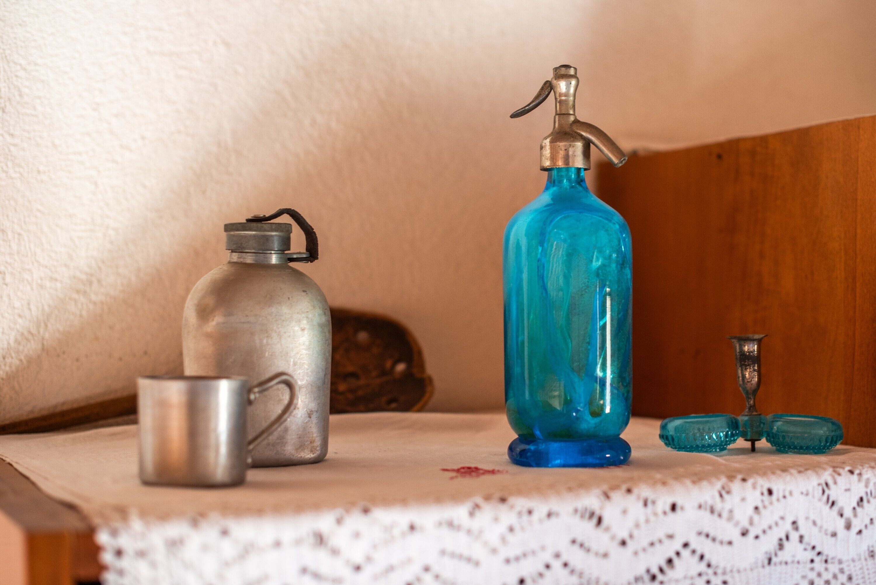 Une bouteille d’eau en métal, une bouteille en verre bleue avec un système de pompe et un gobelet en métal, le tout posé sur une table recouverte d’une nappe en dentelle.