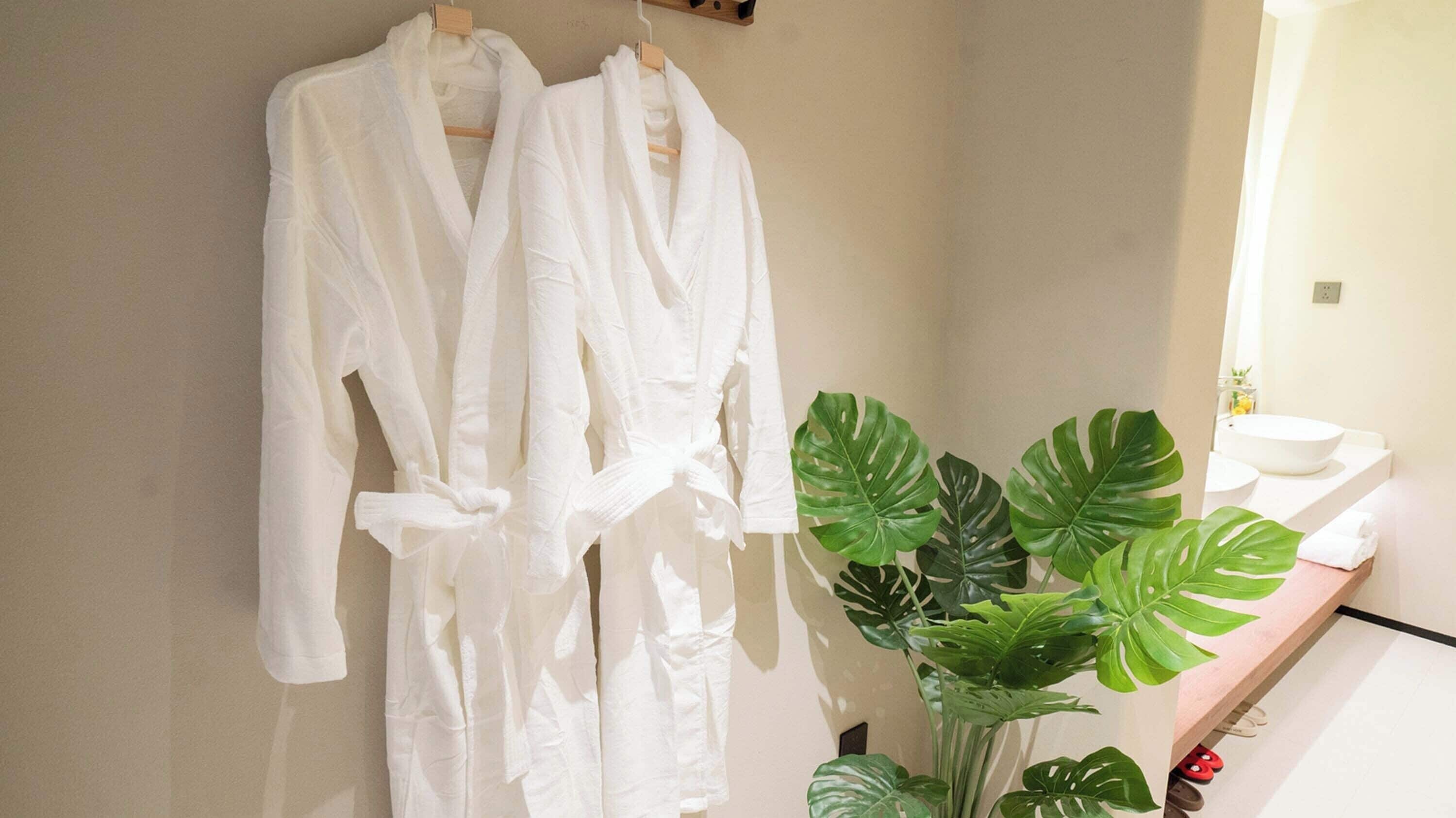 Two white robes hanging on a wooden rack in a bathroom with a sink and a large green plant.