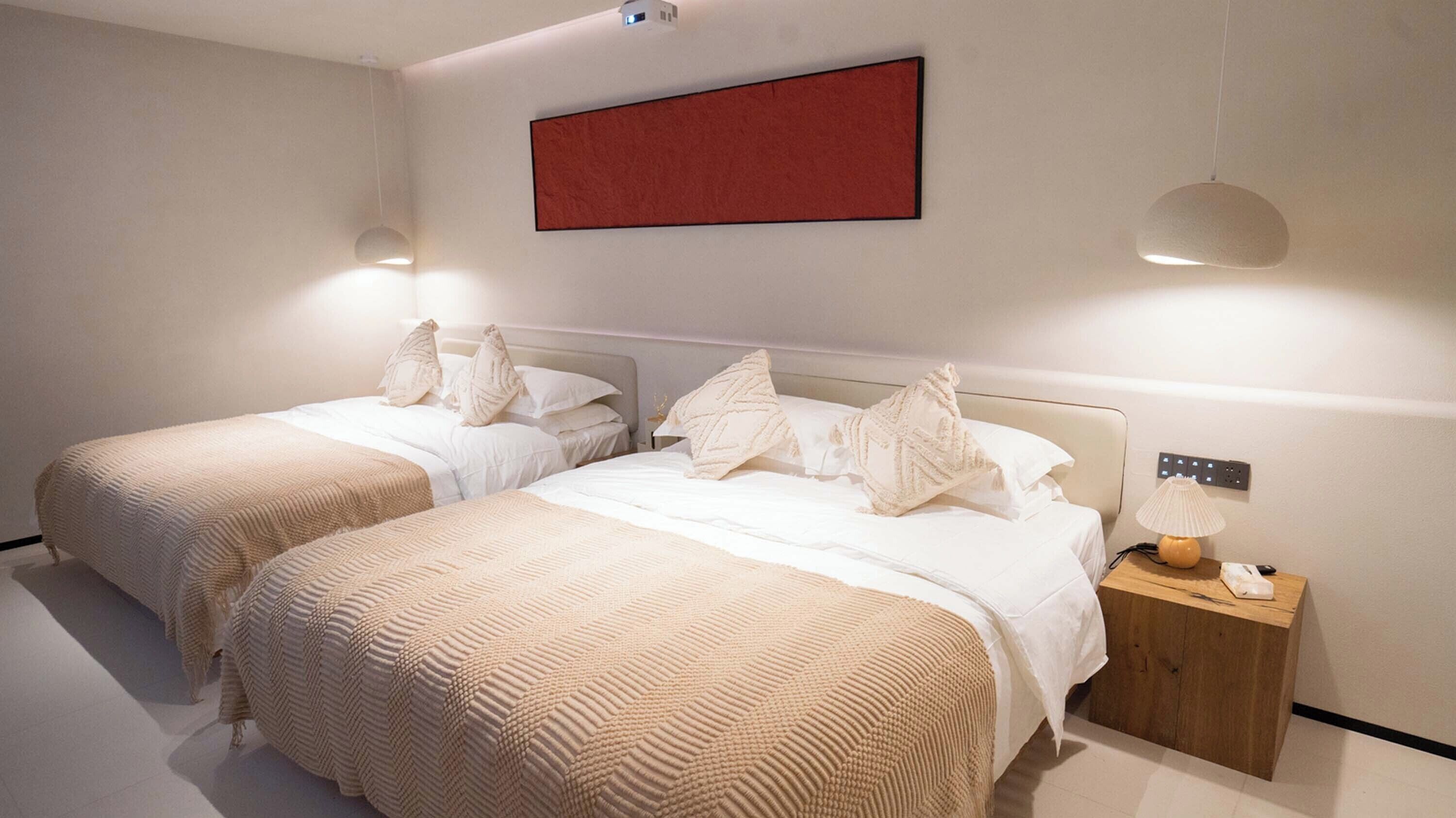 A modern bedroom with two beds, each with a textured beige bedspread and white pillows. There is a red abstract painting on the wall, a pendant light, and a bedside table with a lamp.