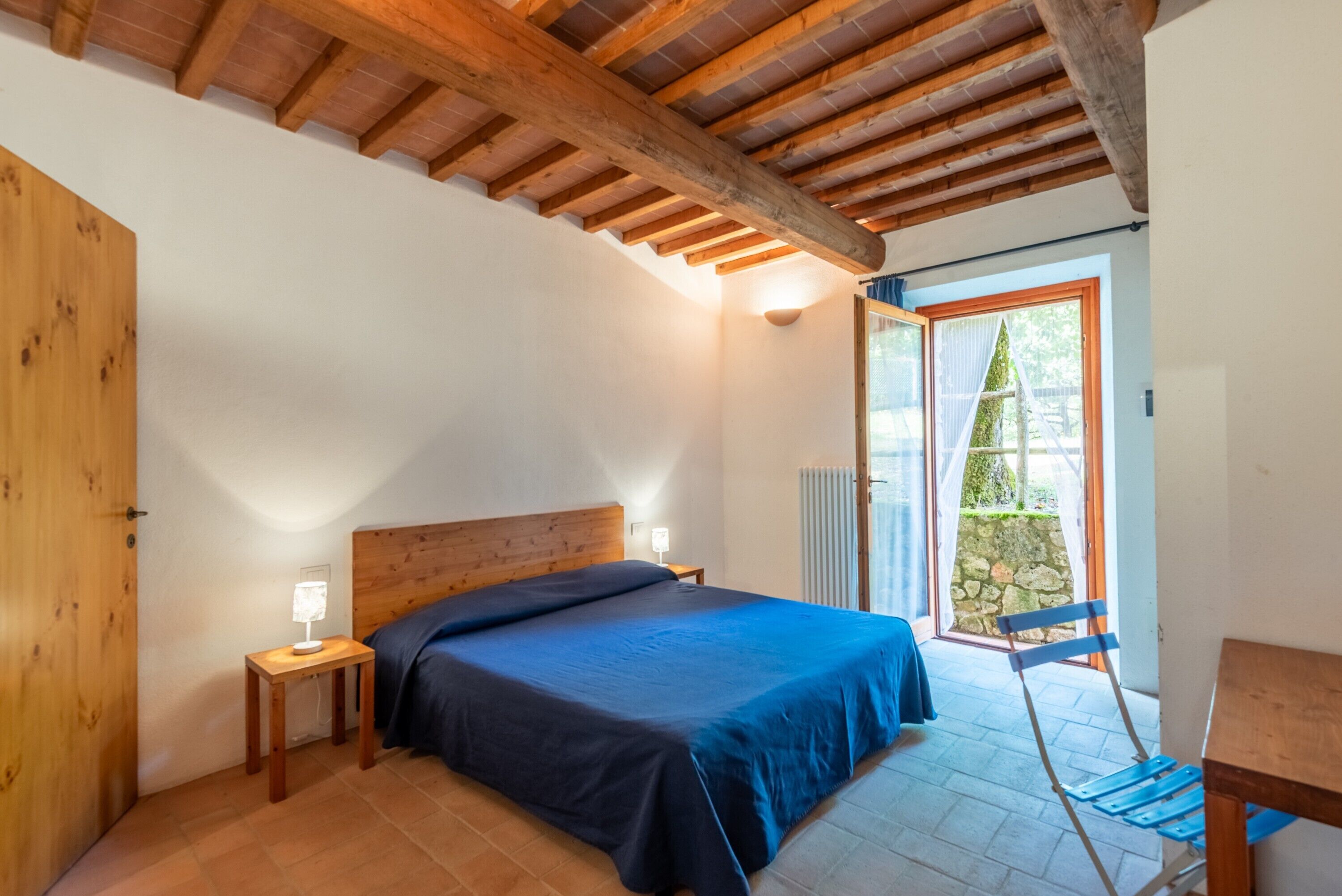 A bedroom with a wooden ceiling, a bed with a blue bedspread, a nightstand with a lamp, and a view of the outside through a glass door.