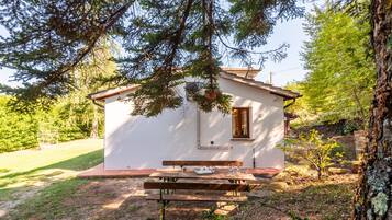 Ein kleines, weißes Blockhaus mit einer hölzernen Bank und einem Picknicktisch davor, umgeben von Bäumen und einer Wiese.