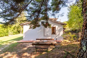 A small white cabin with a wooden bench and picnic table in front, surrounded by trees and a grassy area.