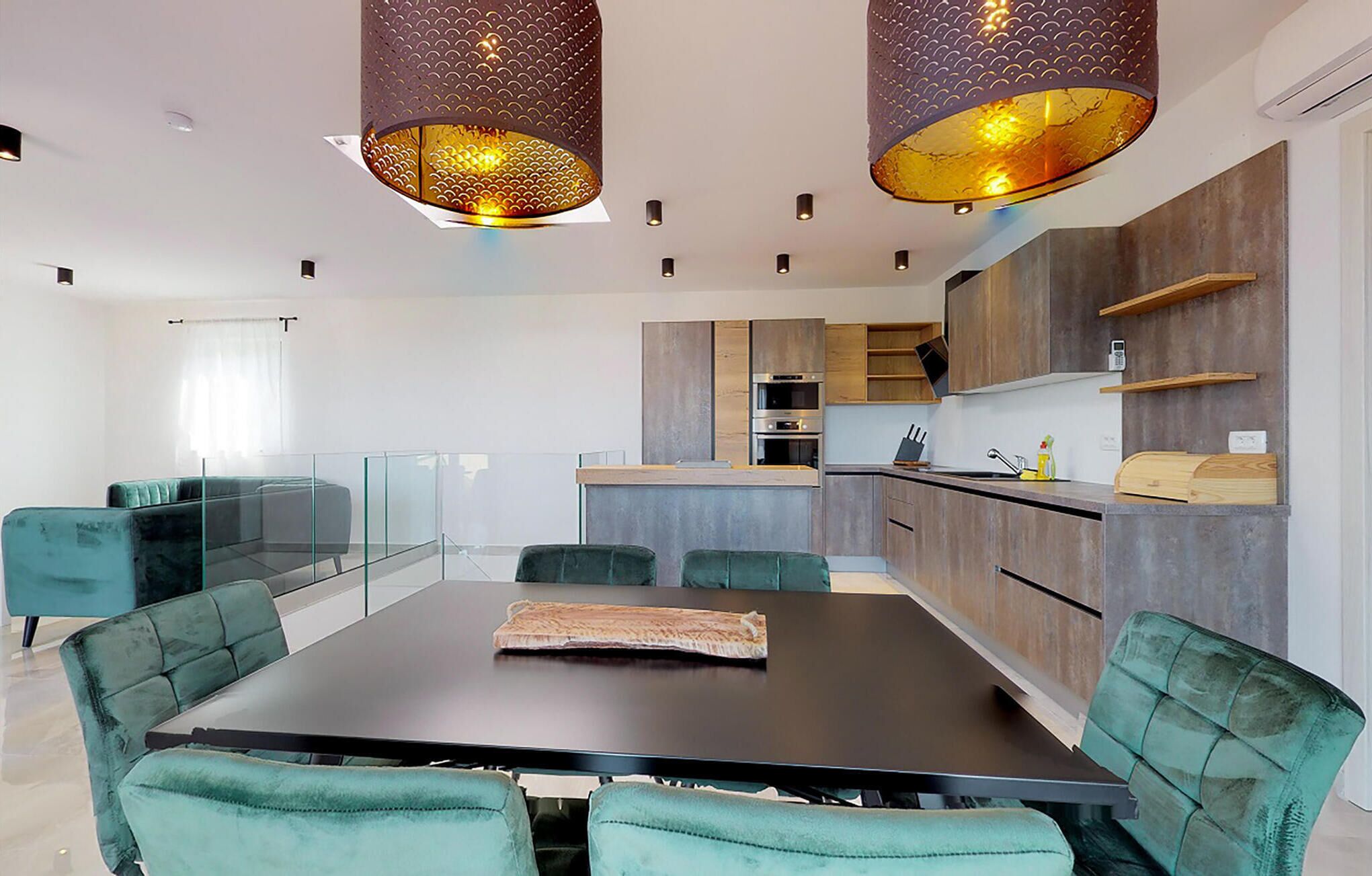 A modern kitchen with a central island, built-in appliances, and a dining area with a glass partition and teal upholstered chairs.