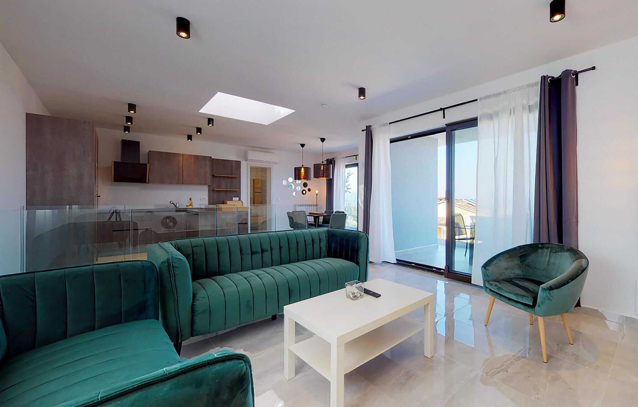 A modern living room with a green sofa, a white coffee table, and a green armchair. There is a glass partition separating the living room from the kitchen area.