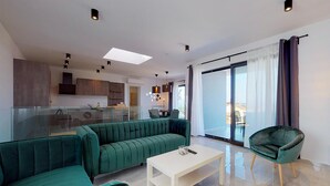 A modern living room with a green sofa, a white coffee table, and a green armchair. There is a glass partition separating the living room from the kitchen area.