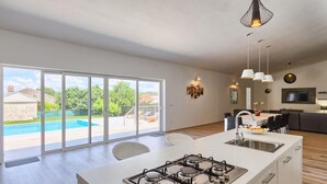 A modern kitchen with a gas stove, a dining area with a table and chairs, and a large sliding glass door leading to a pool and garden. - Beautiful home in Rovinjsko selo (Rovinjsko selo)