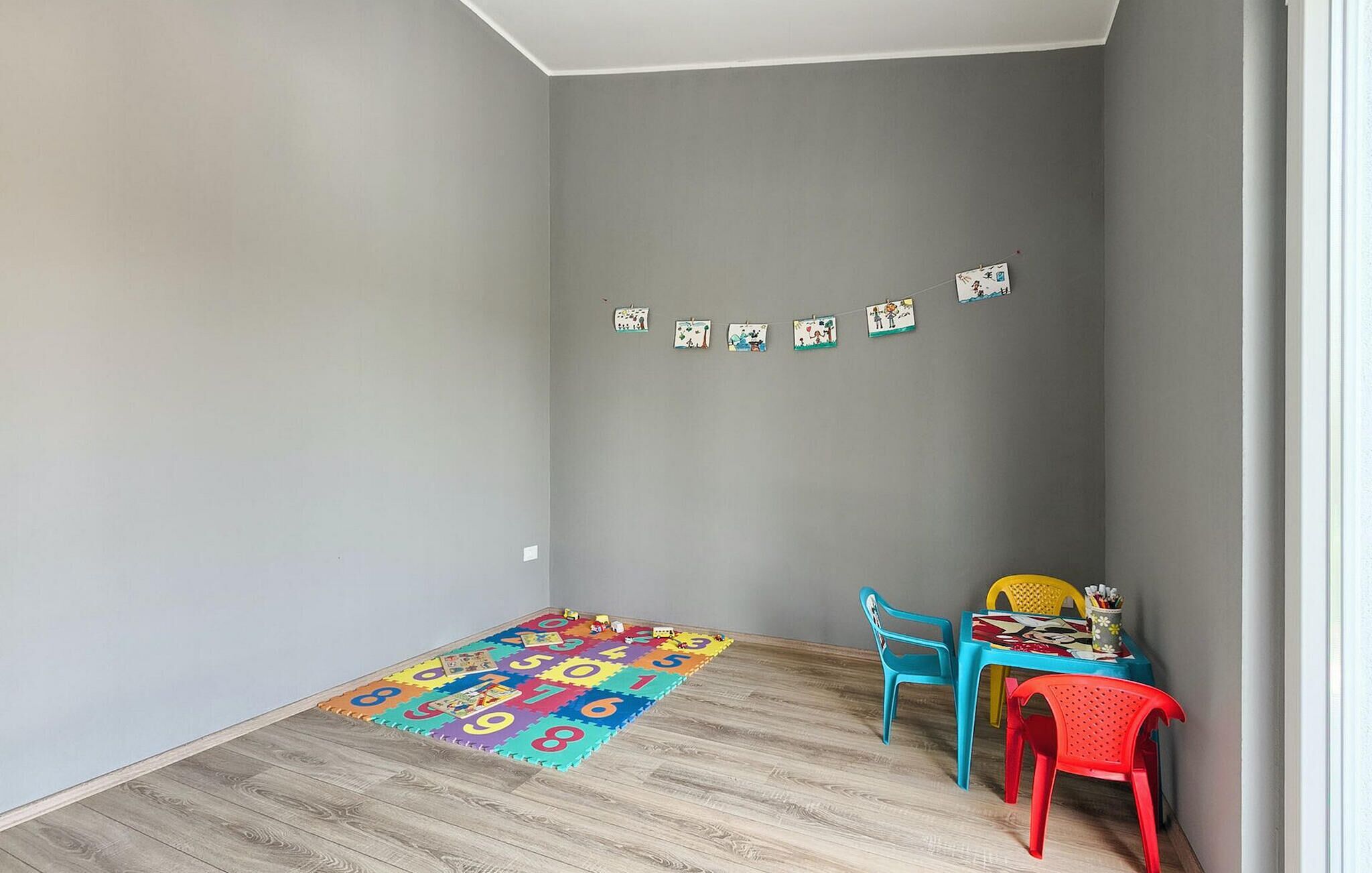 Een kleine kamer met grijze wanden, een houten vloer, een kleurrijk speelkleed en een tafel met stoelen.