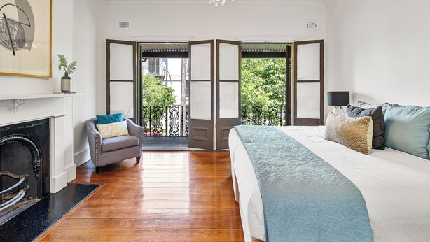 Une chambre avec un grand lit, une cheminée, un fauteuil et une vue sur l’extérieur, visible à travers une porte.