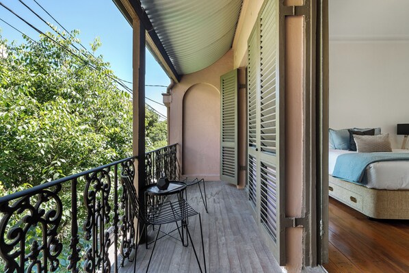A balcony with a table and chairs, a bed with a blue blanket, and a view of trees.