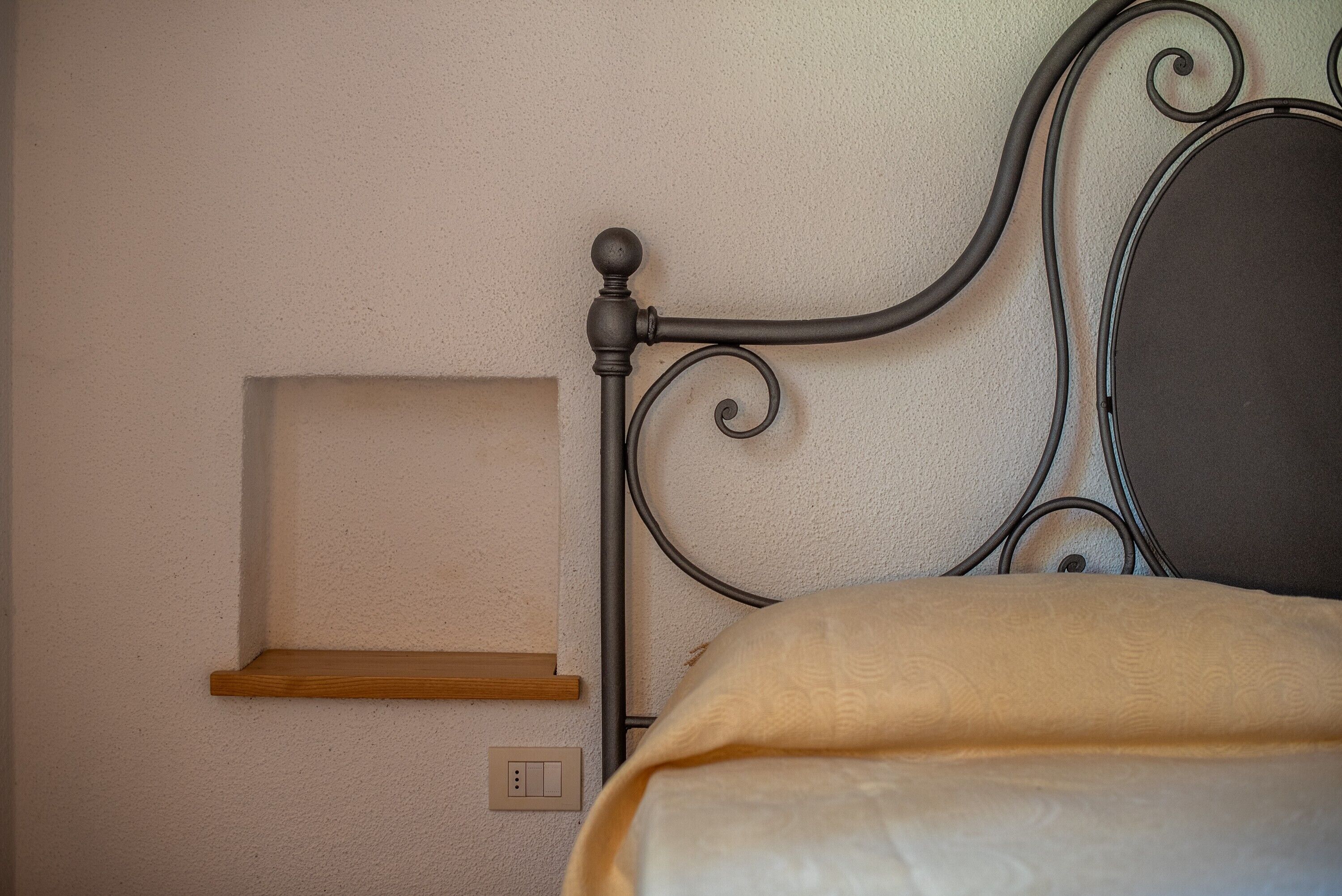A bed with a metal headboard, a wooden shelf, and a light switch on the wall.