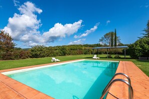 Una piscina rectangular con borde de azulejos, rodeada de césped y árboles.