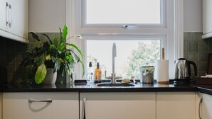 A kitchen with a window, a sink, a potted plant, a bottle of soap, a kettle, and a jar.
