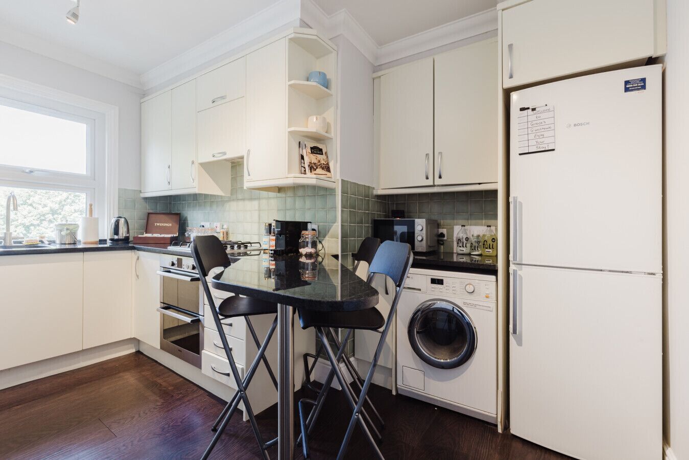Une cuisine moderne avec des meubles blancs, un lave-linge encastré et un coin repas comprenant une table et des chaises en verre.