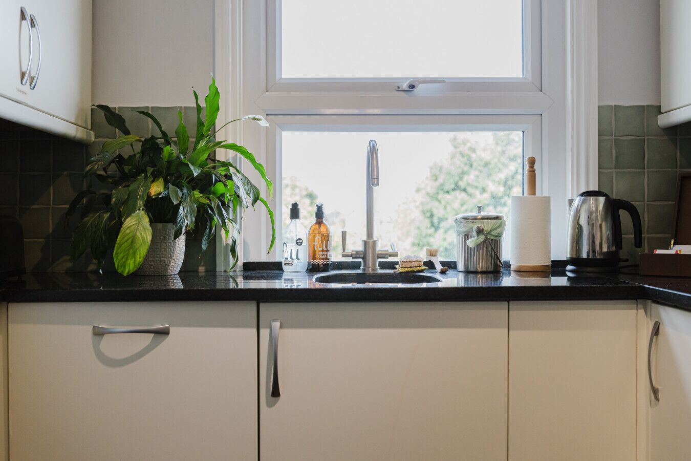 Een keuken met een raam, een gootsteen, een bloempot, een zeepfles, een waterkoker en een pot.