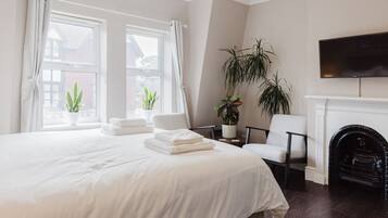 Une chambre d’hôtel avec un grand lit, deux fenêtres avec des rideaux, une plante en pot, une chaise, une cheminée et un téléviseur à écran plat fixé au-dessus de la cheminée.