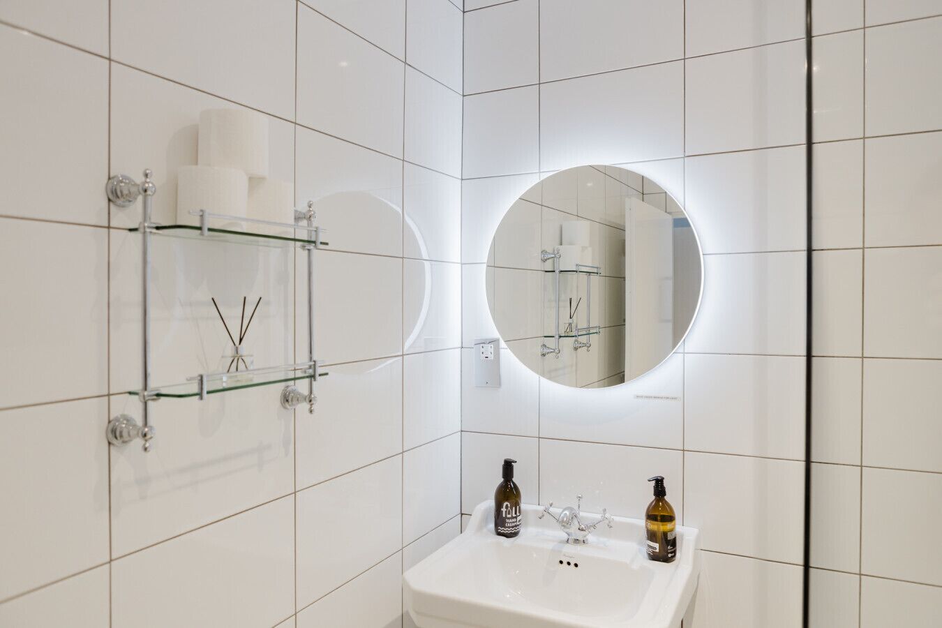 Un baño moderno con azulejos blancos, repisas de vidrio con artículos de aseo, un espejo redondo con iluminación integrada y un lavamanos blanco.
