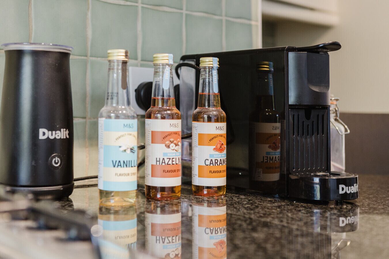 Eine Küchenarbeitsplatte mit verschiedenen Gegenständen, darunter ein Dualit Staubsauger, Flaschen mit M&S Aroma-Sirup und eine Kaffeemaschine.
