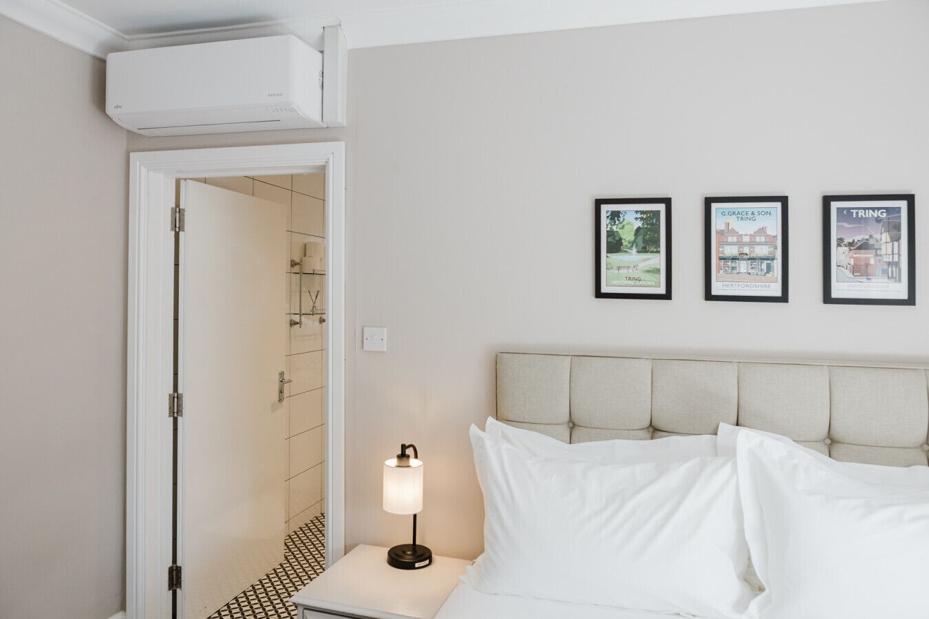 Une chambre d’hôtel avec un lit, une table de chevet avec une lampe, une porte donnant sur une salle de bain et trois tableaux encadrés accrochés au mur.