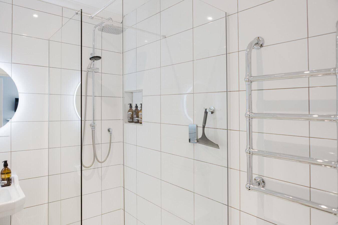 A modern bathroom with white tiles, a glass shower enclosure, a handheld showerhead, a handheld brush, a towel rack, and a mirror.