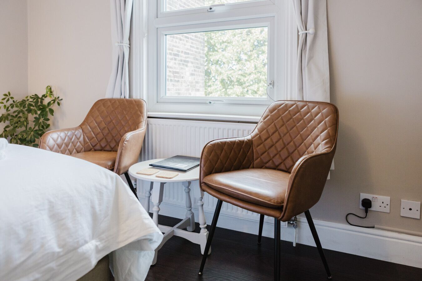 Une pièce comprenant un lit, deux fauteuils en cuir, une petite table ronde avec un livre dessus, et une fenêtre avec des rideaux.