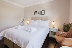 Una cama bien hecha con sábanas blancas, una mesita de noche con lámpara, una silla y un cuadro colgado en la pared.