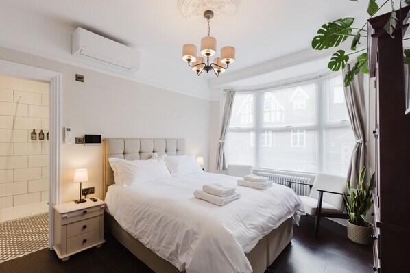 Une chambre d’hôtel avec un grand lit, des tables de chevet, une petite table, un climatiseur fixé au mur, un luminaire au plafond, une fenêtre avec des rideaux et une plante en pot.