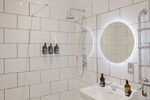 Une salle de bain moderne avec des carreaux blancs, un miroir rond, une douche et trois flacons de produits de soins capillaires Folli accrochés au mur.